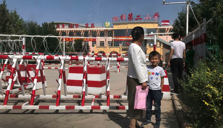 In this Aug. 31, 2018, photo, shot through a car window, a child and a woman wait outside a school entrance with multiple layers of barbed wire and barricades in Peyzawat, western China's Xinjiang region. Uighurs fear the Chinese government's expansion of compulsory Mandarin-intensive classes and boarding schools away from home will gradually erode their children's Central Asian ethnic identity and Islamic beliefs.