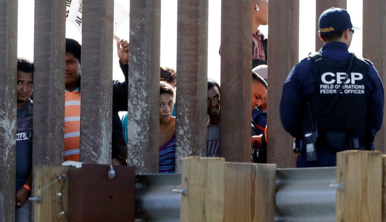 Migrants from Central America look through a border wall as a U.S. Customs and Border Protection officer, right, stands on the other side Sunday, Nov. 25, 2018, seen from San Diego. Migrants approaching the U.S. border from Mexico were enveloped with tear gas Sunday after a few tried to breach the fence separating the two countries.