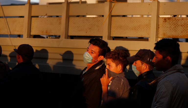 Migrants, their faces covered with masks or a T-shirt as the odor of sewage from overused portable toilets pervades the air, wait in the men's line to receive food from the Mexican Navy outside of a sports complex where more than 5,000 Central Americans are sheltering, Tijuana, Mexico, Wednesday, Nov. 28, 2018.