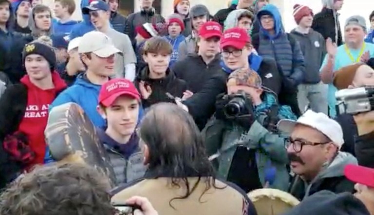 In this Friday, Jan. 18, 2019 image made from video provided by the Survival Media Agency, a teenager wearing a "Make America Great Again" hat, center left, stands in front of an elderly Native American singing and playing a drum in Washington. The Roman Catholic Diocese of Covington in Kentucky is looking into this and other videos that show youths, possibly from the diocese's all-male Covington Catholic High School, mocking Native Americans at a rally in Washington.