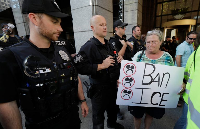 A woman carries a sign that reads "Ban ICE" past a row of law enforcement officers during a protest against family separation at the border and other immigration-related issues, Thursday, Aug. 1, 2019, outside ICE headquarters in Seattle.