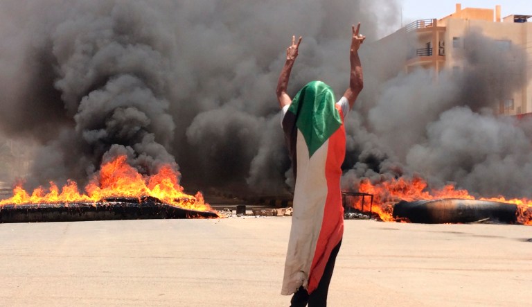 In this June 3, 2019 file photo, a protester wearing a Sudanese flag flashes the victory sign in front of burning tires and debris, near Khartoum's army headquarters, in Khartoum, Sudan. 