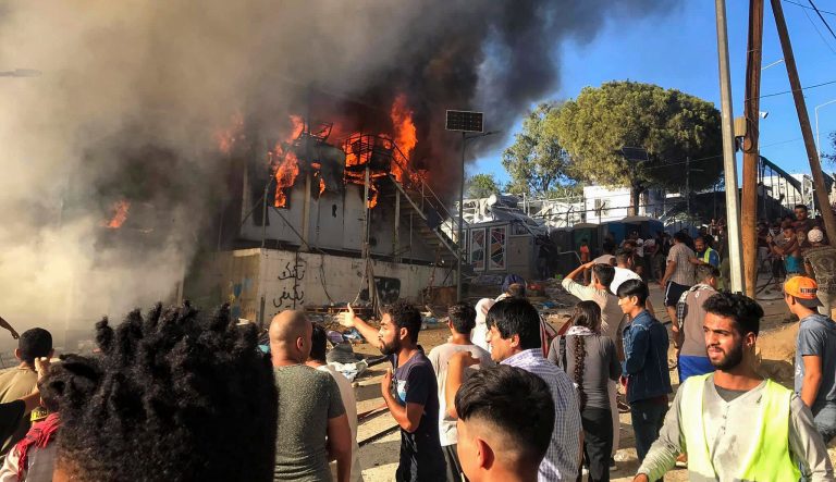 Migrants and refugees stand next to burning house containers at the Moria refugee camp, on the northeastern Aegean island of Lesbos, Greece, Sunday, Sept. 29, 2019.