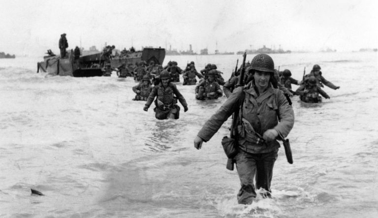 U.S. infantrymen wade through the surf as they land at Normandy in the days following the Allies' June 1944, D-Day invasion of occupied France. An allied ship loaded with supplies and reinforcements waits on the horizon.  