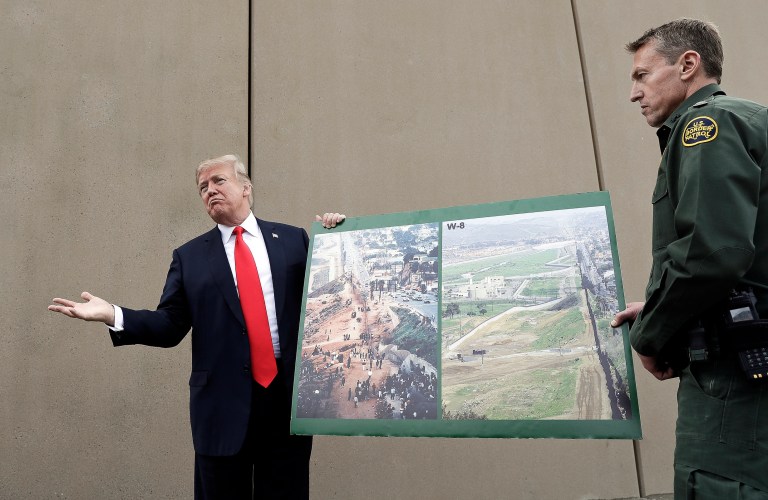 President Donald Trump holds a poster with photographs of the U.S. - Mexico border area as he reviews border wall prototypes in 2018 in San Diego with Rodney Scott, the new head of the U.S. Border Patrol.
