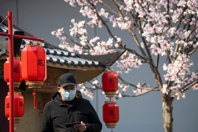 A man wearing a mask looks out from a decoration promoting a movie in Beijing, China on Wednesday, Feb. 19, 2020. U.S. officials are doubling down on putting the blame for the pandemic on Chinese secrecy.