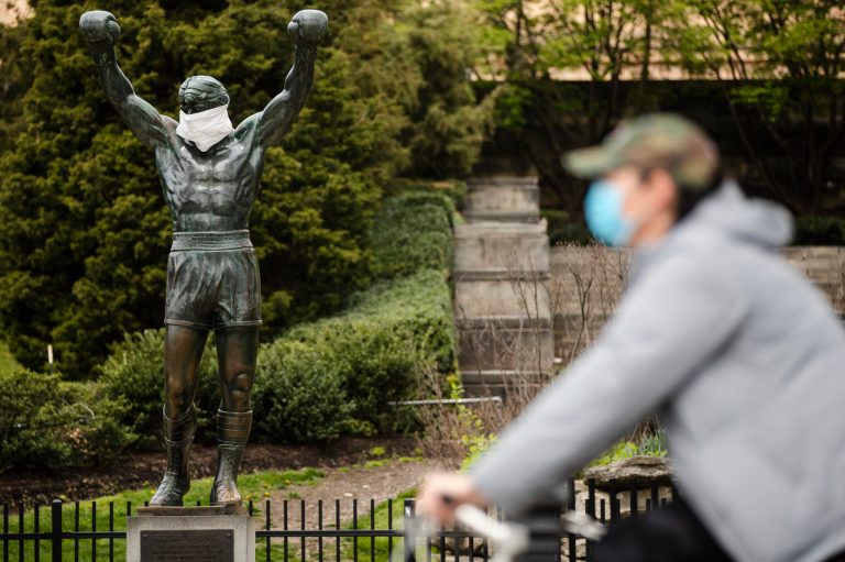 A cyclist wearing a protective face mask as a precaution against the coronavirus moves past the Rocky statue outfitted with mock surgical face mask at the Philadelphia Art Museum in Philadelphia, Tuesday.