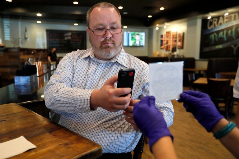 Chris Teague, a customer at CraftWay Kitchen restaurant, scans a QR Code using his smartphone that is on a placard held by employee Laura Ramirez, right, in Plano, Texas, Friday, May 1, 2020. Scanning the code pulls up the restaurants full menu allowing customers to order without handling a paper menu. (AP Photo/Tony Gutierrez)