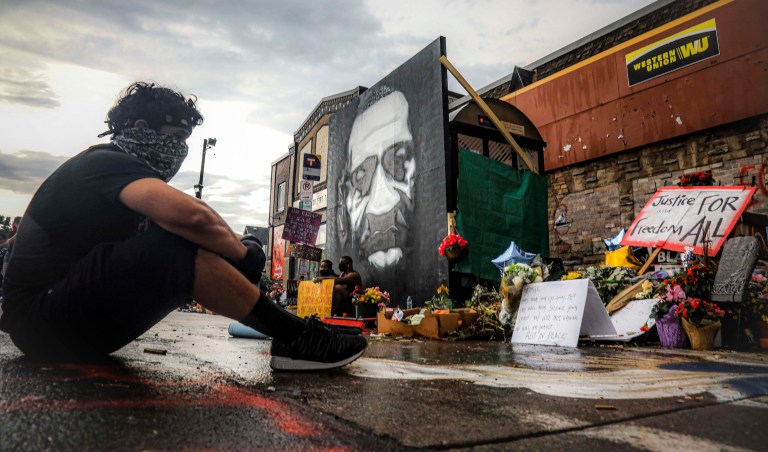After a new mural, center, of George Floyd is added to a growing memorial of tributes, Trevor Rodriquez sits alone at the spot where Floyd died while in police custody, Tuesday June 2, 2020, in Minneapolis, Minn.  âI have been out every single night protesting peacefully, just trying to support everything,â said Rodriquez. âI didnât want to come here just on a rush, so I had to just take a moment to pay my respect.â 
