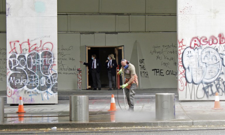 In this July 8, 2020, file photo, a worker washes graffiti off the sidewalk in front of the Mark O. Hatfield Federal Courthouse in downtown Portland, Ore., as two agents with the U.S. Marshals Service emerge from the boarded-up main entrance to examine the damage. Oregonâs largest city is in crisis as violent protests have wracked downtown for weeks. The mayor canât bring the council along on cracking down because of very liberal council members and no matter what he does, the city gets sued anyway. (AP Photo/Gillian Flaccus, File)
