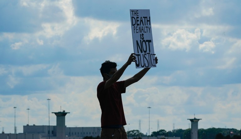 A death penalty protestor carries a sign  across the street from the federal prison complex in Terre Haute, Ind., Wednesday, Aug. 26, 2020.
