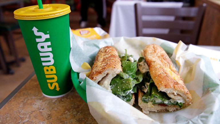 The Subway logo is seen on a soft drink cup next to a sandwich at a restaurant.