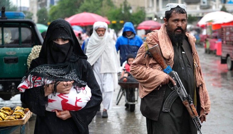 An Afghan woman walks through the old market as a Taliban fighter stands guard, in downtown Kabul, Afghanistan, Tuesday, May 3, 2022.