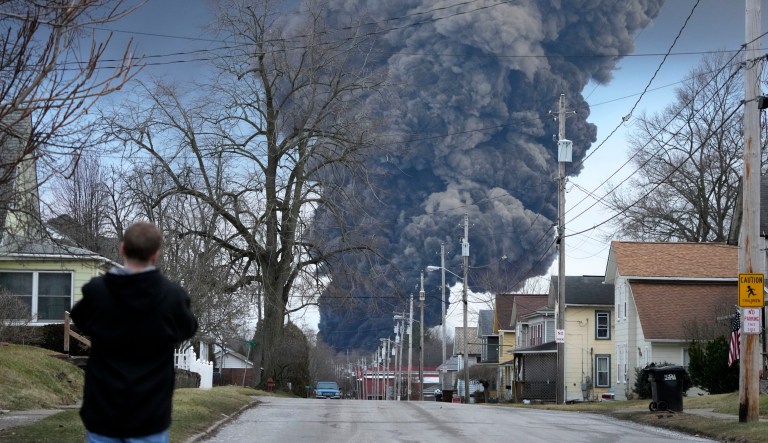 A man takes photos as a black plume rises over East Palestine, Ohio, as a result of a controlled detonation of a portion of the derailed Norfolk Southern train, Feb. 6, 2023. After toxic chemicals were released into the air from a wrecked train in Ohio, evacuated residents remain in the dark about what toxic substances are lingering in their vacated neighborhoods while they await approval to return home.