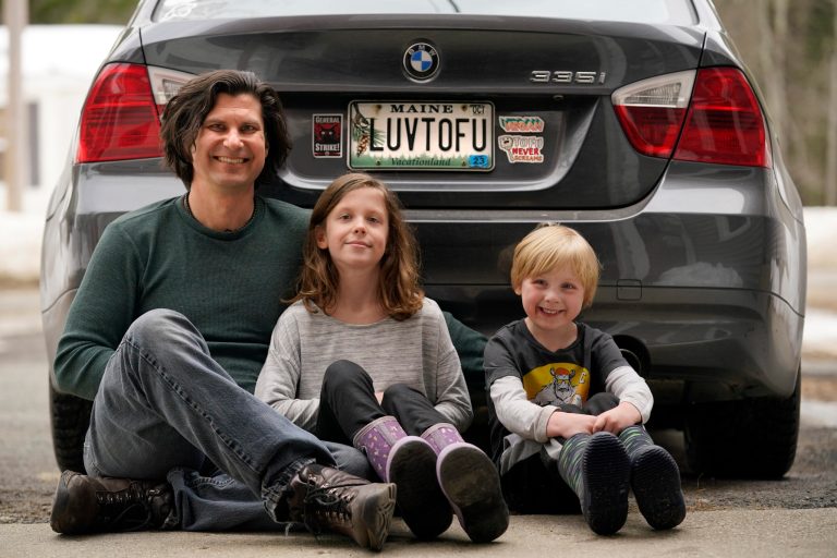 Peter Starostecki and his kids, Sadie, center, and Jo Jo, pose behind their car with the vanity license plate that the state of Maine has deemed inappropriate, March 8, 2023, in Poland, Maine. The vegan family's car will soon have a randomly selected plate.