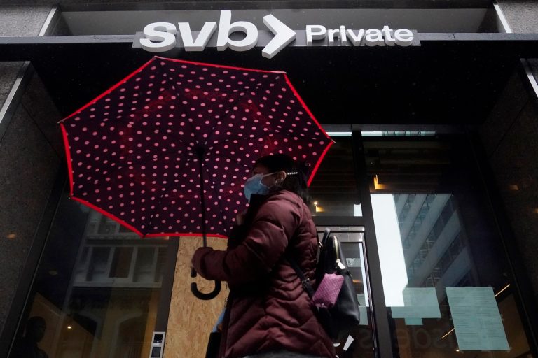 A pedestrian carries an umbrella while walking past a Silicon Valley Bank Private branch in San Francisco, Tuesday, March 14, 2023. After a frenetic weekend of round-the-clock briefings, U.S. policymakers took the audacious step guaranteeing all the deposits of the failed Silicon Valley Banks, even those exceeding the FDIC's $250,000 limit. 