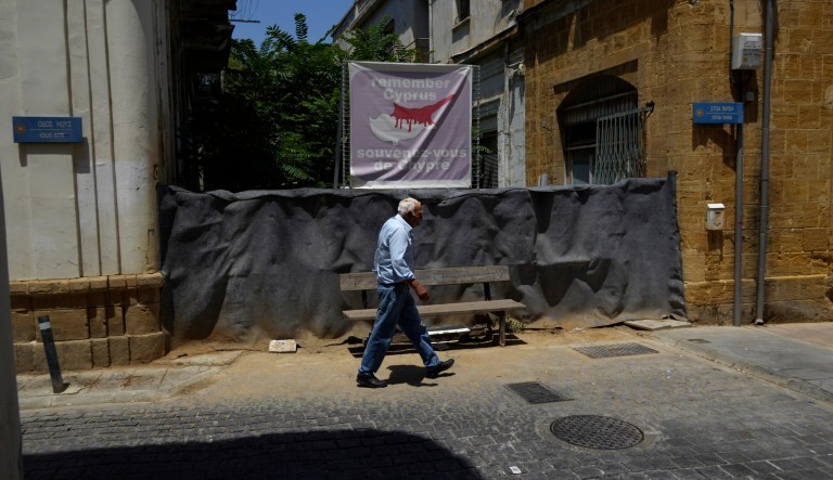 An elderly Cypriot man walks in front of a blocked road with barrels and sand bags where covered with a fabric as on the top is seen a banner showing the island in division, across the UN buffer zone that divides the Greek, south, and Turkish Cypriots, north, controlled areas, in the divided capital Nicosia, in the eastern Mediterranean island of Cyprus, on Wednesday, July 5, 2023. The island's division came about in 1974 when Turkey invaded in the wake of a coup aimed at union with Greece.