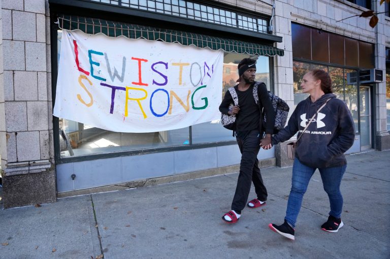 A couple walks by a banner that was put up in response to this week's deadly mass shootings, Friday, Oct. 26, 2023, in Lewiston, Maine. Police are still searching for the suspect who killed at least 18 in separate shootings at a bowling alley and restaurant on Wednesday. 