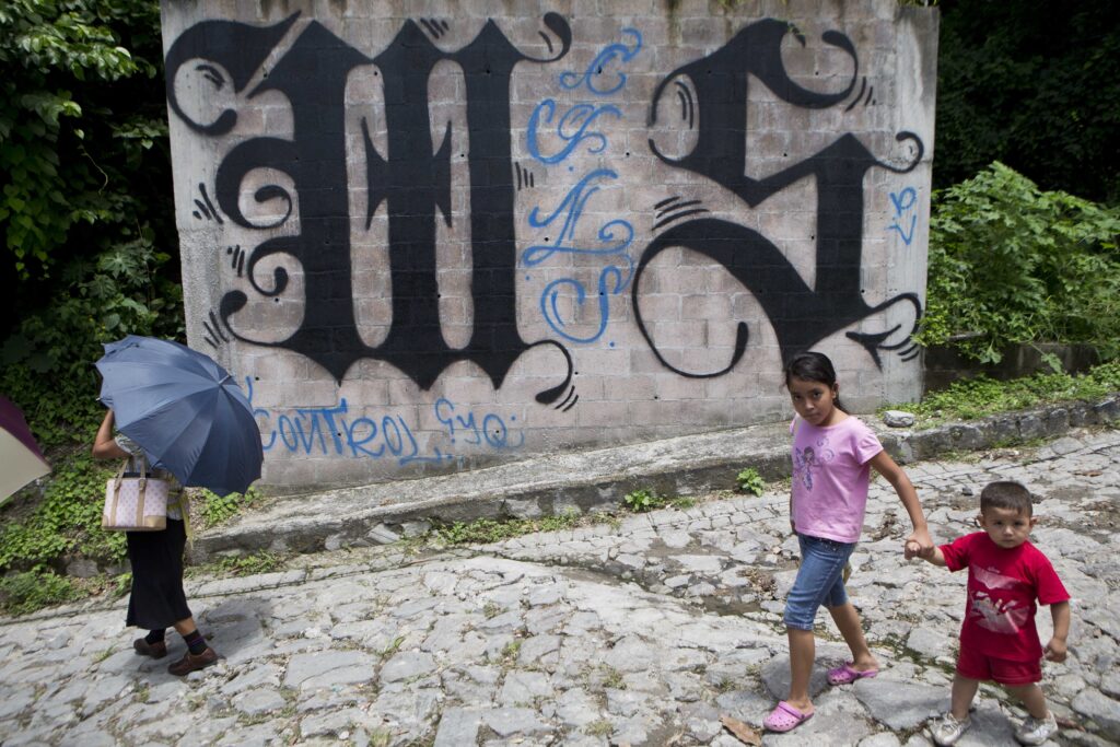 ADVANCE FOR SUNDAY, SEPT. 7 AND THEREAFTER - In this Thursday, Aug. 21, 2014 photo, a family walks by a wall covered by a symbol from the Mara Salvatrucha gang in Ilopango, El Salvador. In Ilopango and communities across El Salvador, the Mara Salvatrucha and their arch rivals, the 18th Street Gang, are de facto rulers. (AP Photo/Esteban Felix)