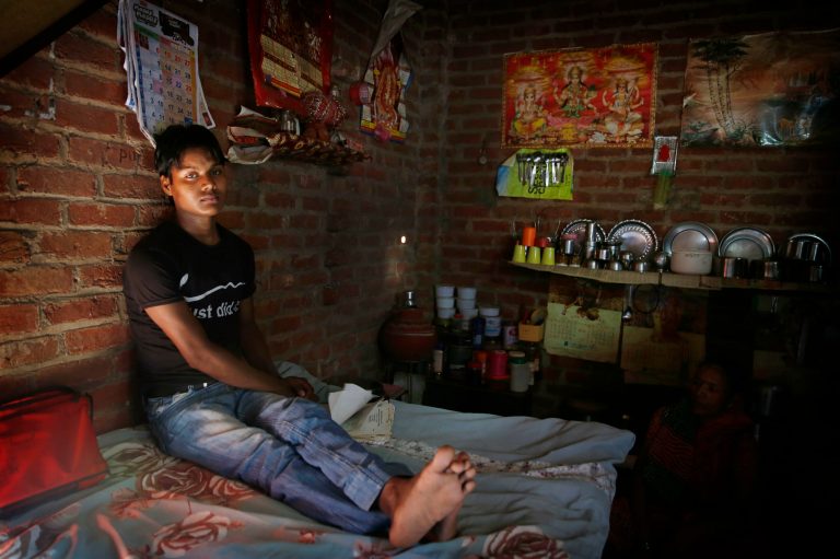 In this July 25, 2014 photo, Digamber Rawat sits in his one room house in New Delhi, India. Rawat rarely emerges from the tiny windowless room he shares with his parents because a mysterious illness has wasted away the strength in his legs. His family can't afford private health care, but they must pay for it anyway, even when they go to free government hospitals for help. (AP Photo/Manish Swarup)