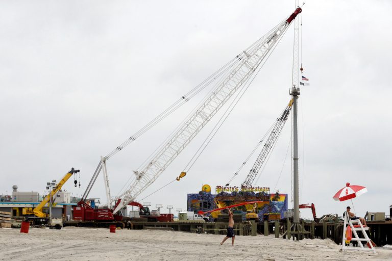 In this Friday, May 24, 2013 photo, Ryan Murphy, center, carries a surfboard and Kenny Dougherty stands guard on a lifeguard stand near the Casino Pier which was ripped apart by Superstorm Sandy, in Seaside Heights, N.J. The Jersey Shore beaches officially opened for the summer on Friday, after rebuilding following the destruction left behind by Superstorm Sandy last fall. The storm caused $37 billion of damage in the state. (AP Photo/Julio Cortez)