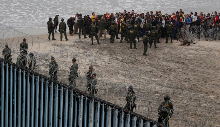 U.S. police and Border Patrol hold a line as members of an inter-faith group, showing support for Central American asylum-seekers who arrived in recent caravans and calling for an end to detaining and deporting immigrants, pray during a protest in San Diego, as seen from across the border wall in Tijuana, Mexico, Monday, Dec. 10, 2018.