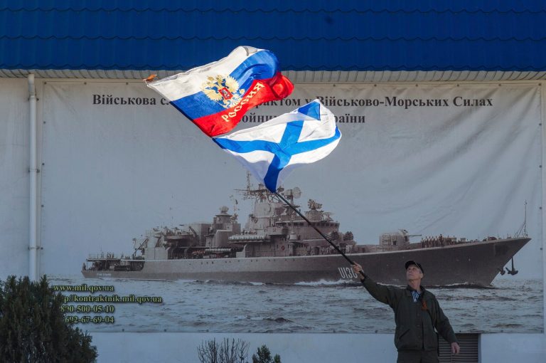 A pro-Russian activist waves the Russian state, upper, and Russian Navy flags outside an entrance to the General Staff Headquarters of the Ukrainian Navy in Sevastopol, Ukraine, Monday, March 3, 2014. Pro-Russian soldiers seem to further cement their control over the strategic region -- that also houses the Russian Black Sea Fleet -- by seizing a ferry terminal in the Ukrainian city of Kerch about 20 kilometers (12 miles) by boat to Russia, intensifying fears that Moscow will send even more troops into the peninsula. It comes as the U.S. and European governments are trying to figure out ways to halt and reverse the Russian incursion. (AP Photo/Andrew Lubimov)