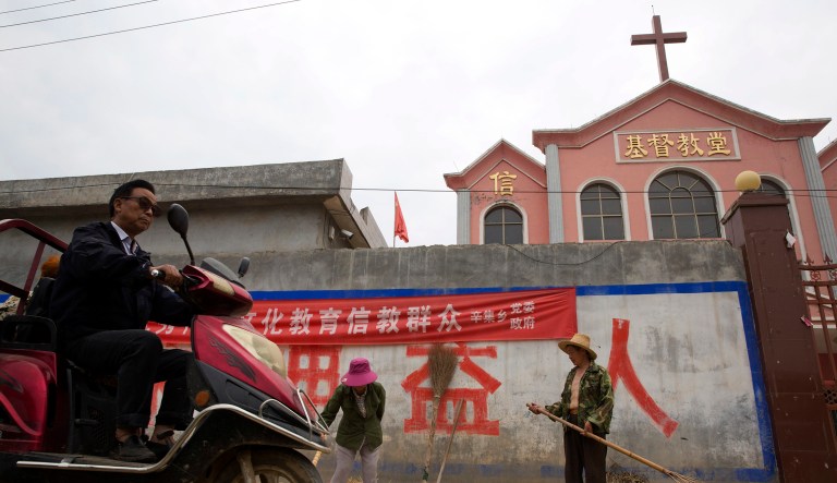 In this photo taken Saturday, June 2, 2018, a man rides past workers tossing hay outside a church with part of a slogan that reads "Educate the believers with excellent Chinese traditional culture" near the city of Pingdingshan in central China's Henan province.