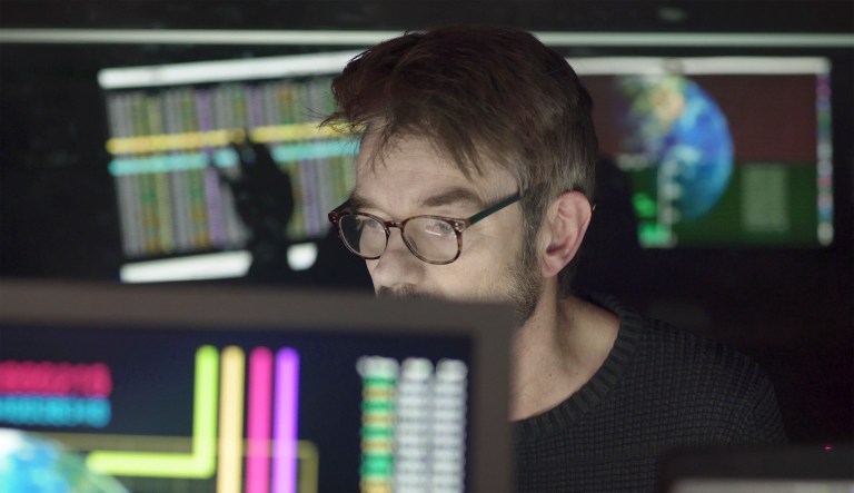 A bearded man studying computer displays depicting global trends with figures, the screens being reflected in his glasses as well as behind him on a black glass wall. 