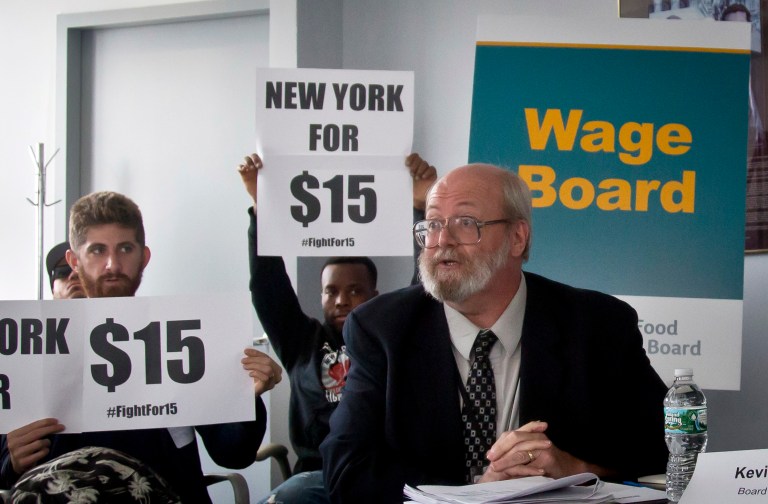 James Brown, principal economist for the New York State Department of Labor, speaks during the first meeting of the state wage board, May 20, 2015, in New York, as demonstrators hold up signs for $15 an hour pay. The panel, established by Gov. Andrew Cuomo, is considering raising the minimum wage for those in the fast food industry. New York's current minimum wage is $8.75 an hour (AP Photo/Bebeto Matthews)