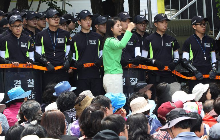 An Evangelical Baptist Church believer shouts slogans against the government as police officers stand guard in font of believers sitting by the main gate of the church in Anseong, South Korea, Wednesday, June 11, 2014. Thousands of South Korean police officers stormed a church compound Wednesday in their hunt for a fugitive billionaire businessman over April's ferry sinking that left more than 300 people dead or missing, officials said. (AP Photo/Ahn Youn-joon)