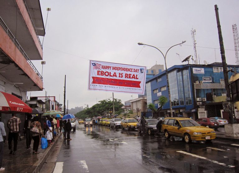 In this photo taken on Monday, July 28, 2014, people hang out in a street under a  banner which warns people to be cautious about Ebola, in Monrovia, Liberia. Two American aid workers in Liberia have tested positive for the virus and are being treated there. U.S. health officials said Monday that the risk of the deadly germ spreading to the United States is remote. (AP Photo/Jonathan Paye-Layleh)