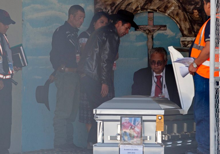 This photo taken through a chain-linked fence, shows Francisco Ramos, wearing a black hat, surrounded by immigration officials, as he identifies the body of his son, whose decomposed body was found in the Texas desert, in Guatemala City, Friday, July 11, 2014. The remains of Gilberto Francisco Ramos , the 15 year old boy who died in the Rio Grande Valley of South Texas, trying to reach the United States alone, was delivered to his family on Friday. (AP Photo/Moises Castillo)