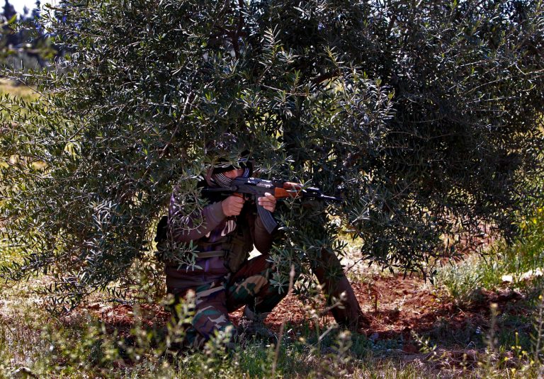 In this Friday, April 12, 2013 photo, a member of the Lebanese pro-Syrian Popular Committees aims his weapon at the Lebanon-Syria border, near the northeastern Lebanese town of al-Qasr, Lebanon. Masked men in camouflage toting Kalashnikov rifles fan out through a dusty olive orchard, part of a group of Hezbollah-backed fighters from Lebanon who are patrolling both sides of a porous border stretch with Syria. The gunmen say their mission to protect Shiites in both countries and counter what they see as a growing threat from Sunni rebels in Syria. (AP Photo/Bilal Hussein)