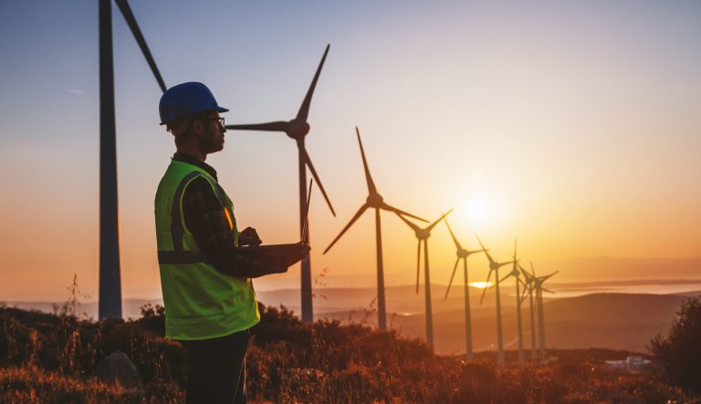 Silhouette of young male engineer holding laptop computer planning and working for the energy industry and standing beside a wind turbines farm power station.