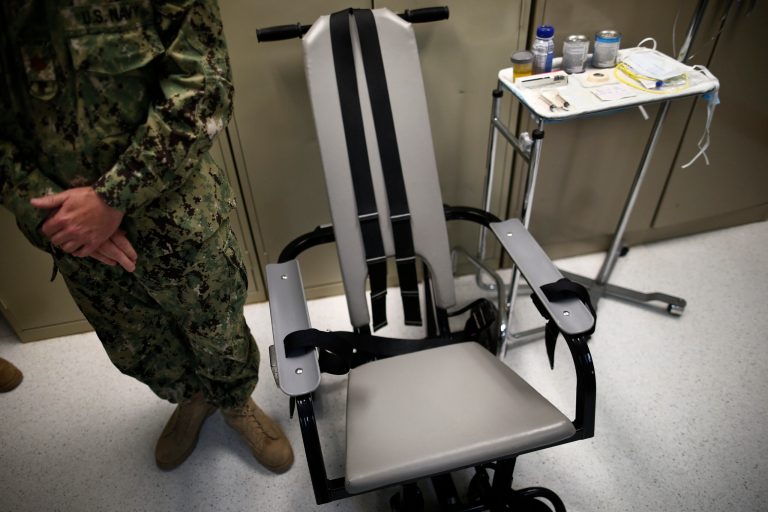 FILE - In this photo Nov. 20, 2013 file photo reviewed by the U.S. military, a U.S. Navy nurse stands next to a chair with restraints, used for force-feeding, and a tray displaying nutritional shakes, a tube for feeding through the nose, and lubricants, including a jar of olive oil, during a tour of the detainee hospital at Guantanamo Bay Naval Base in Cuba. Attorneys for a Syrian prisoner have begun studying hours of video showing him being removed from his cell, placed in a restraint chair and fed by a tube with liquid nutrients. They are looking for evidence of what he has portrayed to them as abusive force-feeding, akin to torture, during the months that he has participated in a hunger strike that drew the attention of President Barack Obama and led to a renewed effort to close the prison. (AP Photo/Charles Dharapak, File)