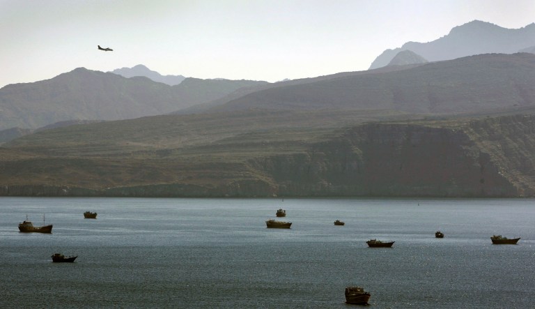 In this Jan. 19, 2012 photo, a plane flies over the mountains in south of the Strait of Hormuz as the trading dhows and ships are docked on the Persian Gulf waters near the town of Khasab, in Oman. Even as sanctions squeeze Iran ever tighter, there's one clandestine route that remains open for business: A short sea corridor connecting a rocky nub of Oman with the Iranian coast about 35 miles (60 kilometers) across the Gulf.