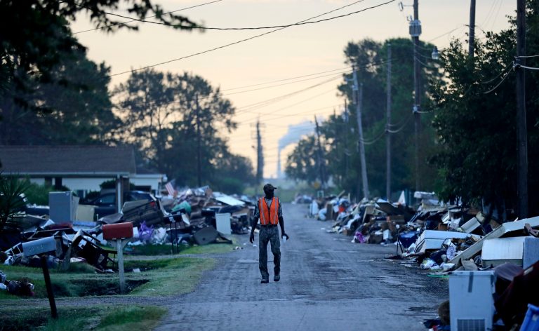 FILE - In this Sept. 28, 2017, file photo, a man walks past debris from homes on his street damaged in flooding from Hurricane Harvey as an oil refinery stands in the background in Port Arthur, Texas. Although many Texas families are still struggling to recover from Hurricane Harvey a year after it caused widespread damage and flooding along the Gulf Coast and in and around Houston, daily life has mostly returned to normal in many of the hardest hit communities.