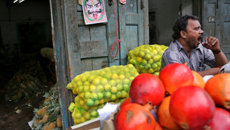 A motif, believed by Hindus to keep evil spirit away, hangs behind as an Indian fruit seller yawns while waiting for buyers at a wholesale market in Bangalore, India, Thursday, Aug. 9, 2018. 