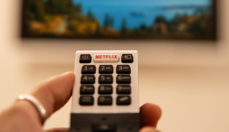 A hand holding television remote control with dedicated Netflix button in front of defocused smart TV. 