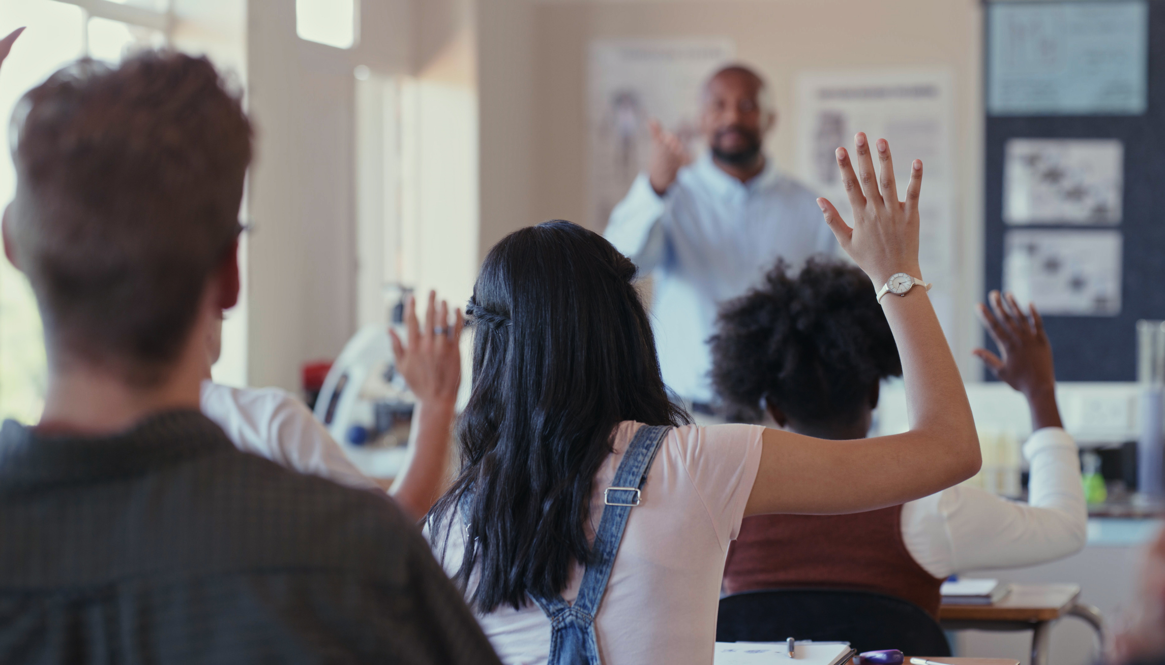 Rearview shot of students raising their hands during a lesson with a teacher in a classroom