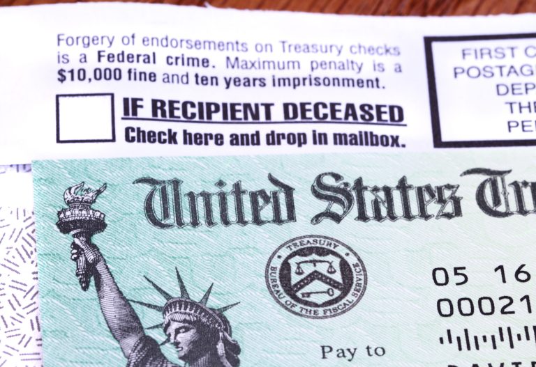 A United States government Treasury Check used to pay Social Security or Medicare benefits lies on top of the opened envelope used to mail it to the recipient. The envelope has warnings of criminal penalties for anyone who fraudulently endorses the check. It also warns that if the recipient is deceased, the check should be placed in the return mail. The distinctive green checks feature an engraving of the Statue of Liberty and the Treasury Seal.