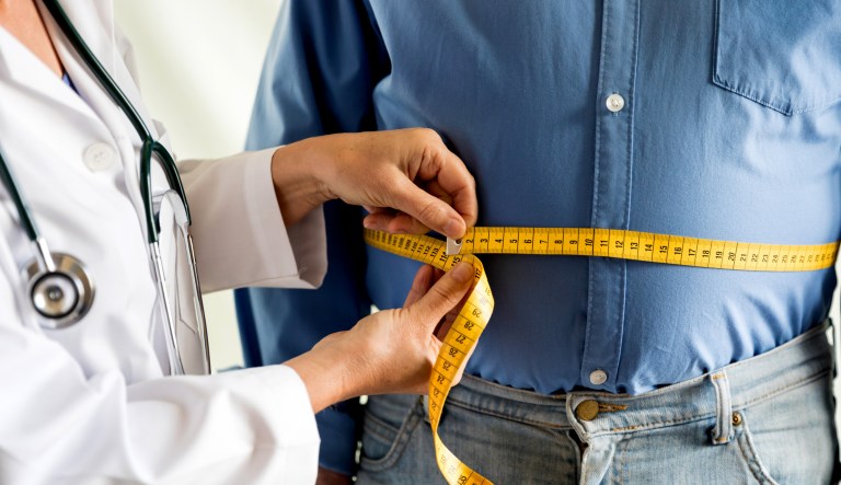 A stock photo shows a doctor with a measuring tape over an overweight man's waist.