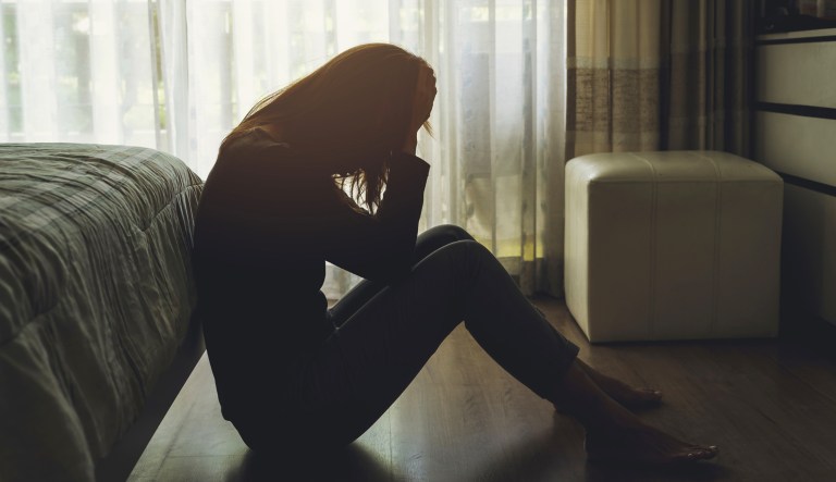 A depressed woman sits in a dark bedroom with her hands covering her face.