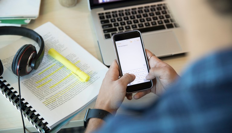 Male college student texting with cell phone, studying at laptop.