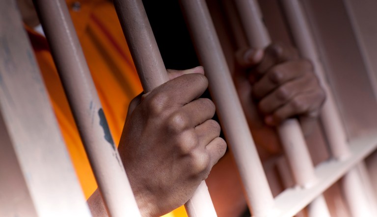A black man with hands outside the bars of a prison cell.