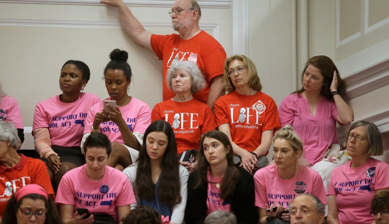 Proponents of a proposed Massachusetts legislative bill, called the "Roe Act" by supporters, wear pink shirts, seated left and right, while opponents of the bill wear red shirts, center top, as they sit near one another in the gallery during a public hearing at the Statehouse, Monday, June 17, 2019, in Boston.