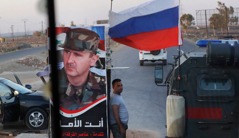 Syrian army soldier stands at a check-point as Russian military police vehicle, right, passes by near the village of Almajdiyeh, Syria, Tuesday, Aug. 14, 2018. The Russian military said Tuesday that its forces in Syria will help U.N. peacekeepers fully restore patrols along the frontier with the Israeli-occupied Golan Heights, reflecting Moscow's deepening role in mediating between the decades-old foes.
