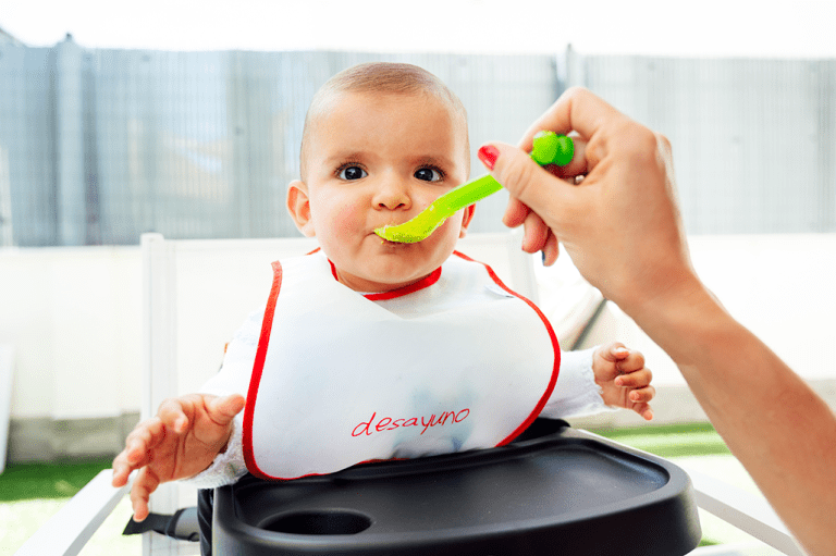 A baby wearing a bib is fed while sitting on a high chair.