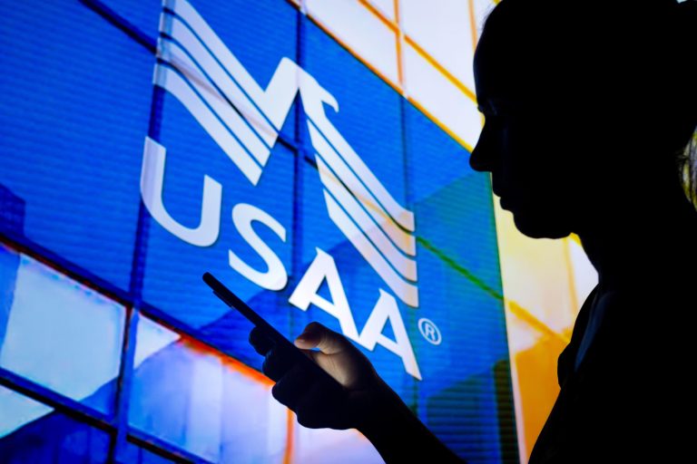 In this photo illustration, a woman's silhouette holds a smartphone with the United Services Automobile Association logo in the background.